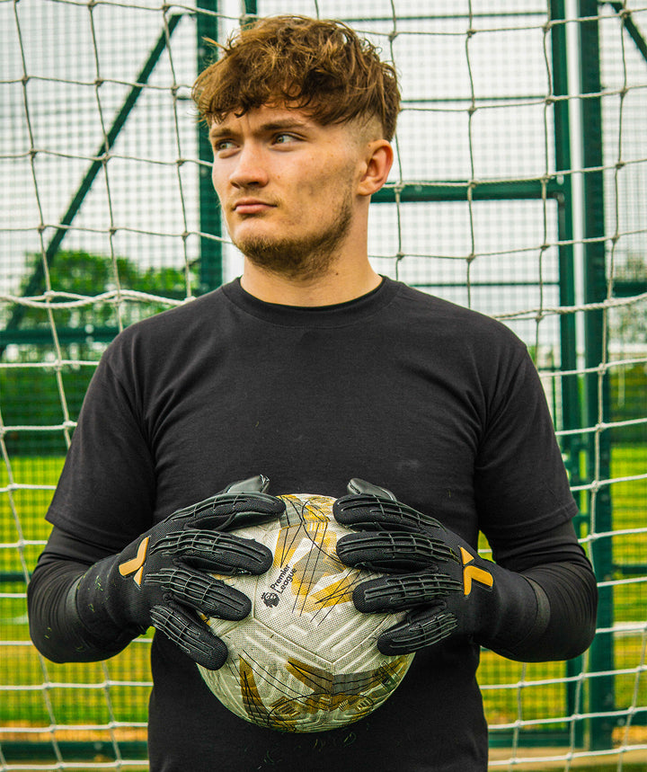 Person wearing black Professional Goalkeeper gloves with a logo, standing in front of a netted background