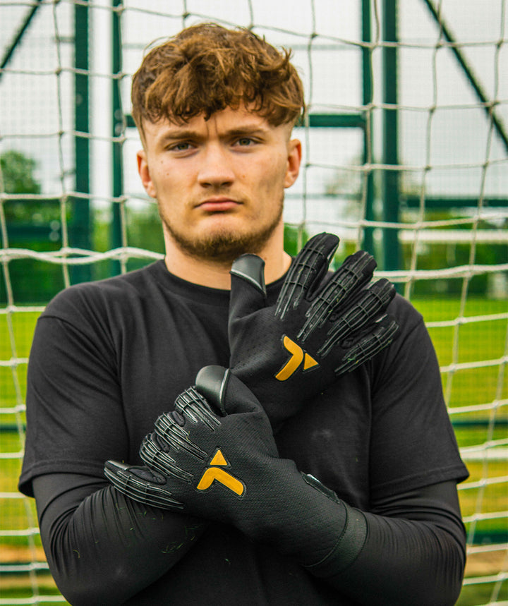 Person wearing black Professional Goalkeeper gloves with a logo, standing in front of a netted background